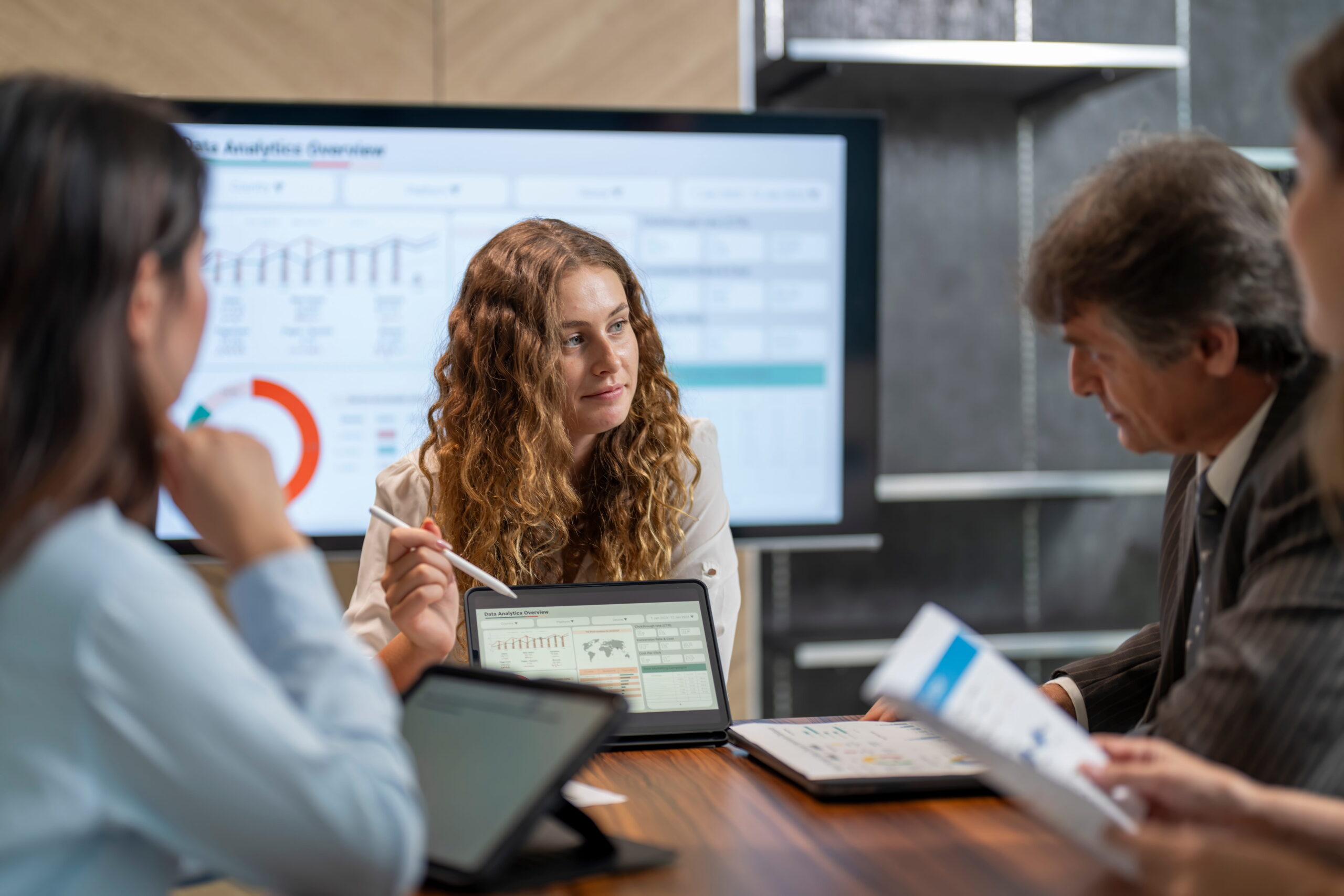 A group of people sitting in a meeting room reviewing documents with a few dashboards on the screen