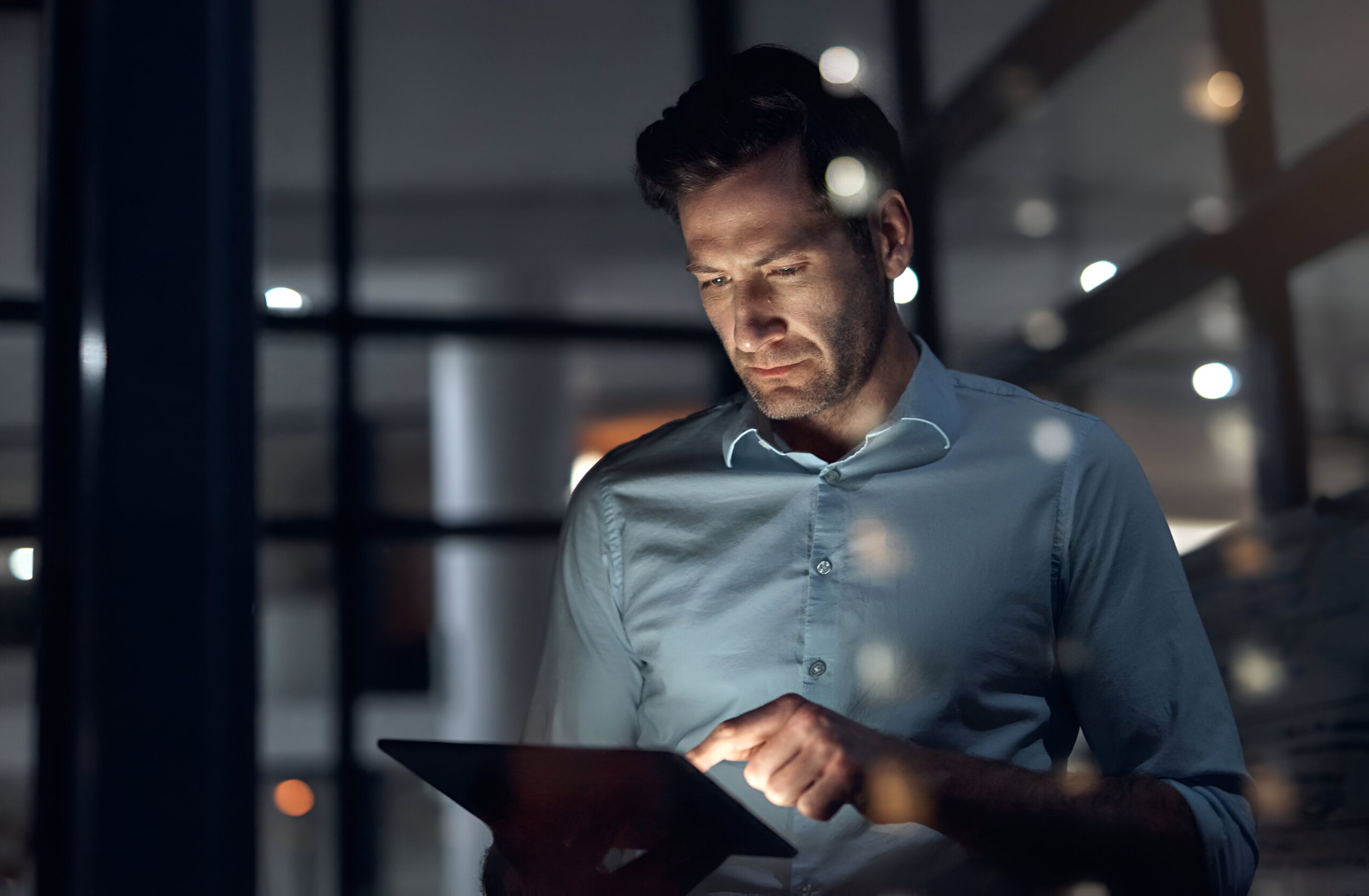 Man in a light blue shirt uses a tablet in a dim office at night, with window frames and blurred reflected lights around him.