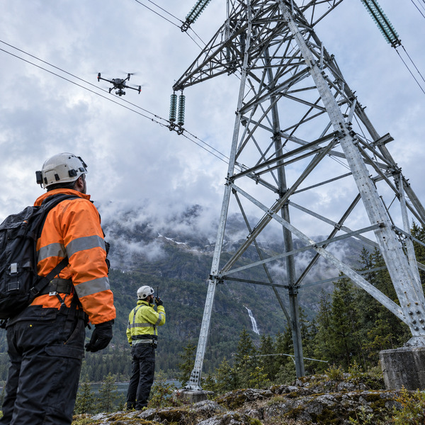 Utility engineers inspect a high-voltage transmission tower as an industrial drone examines insulators in a mountainous corridor.