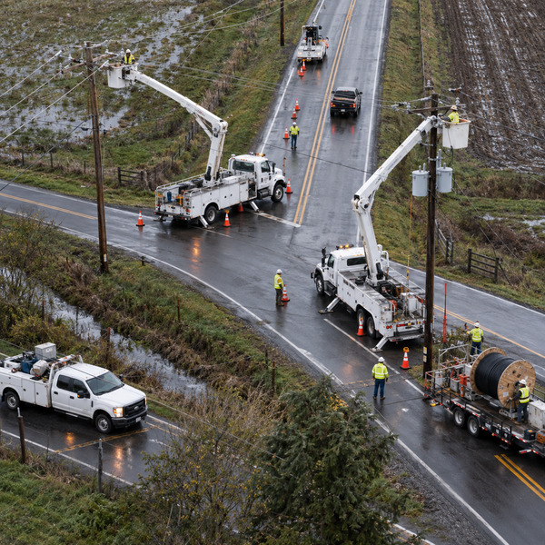 An aerial view shows bucket trucks and ground crews coordinating distribution-line work along a rain-wet rural junction.