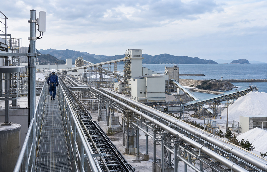 Technician crossing an elevated catwalk above pipelines and distributed salt plant buildings on the Japanese coast.